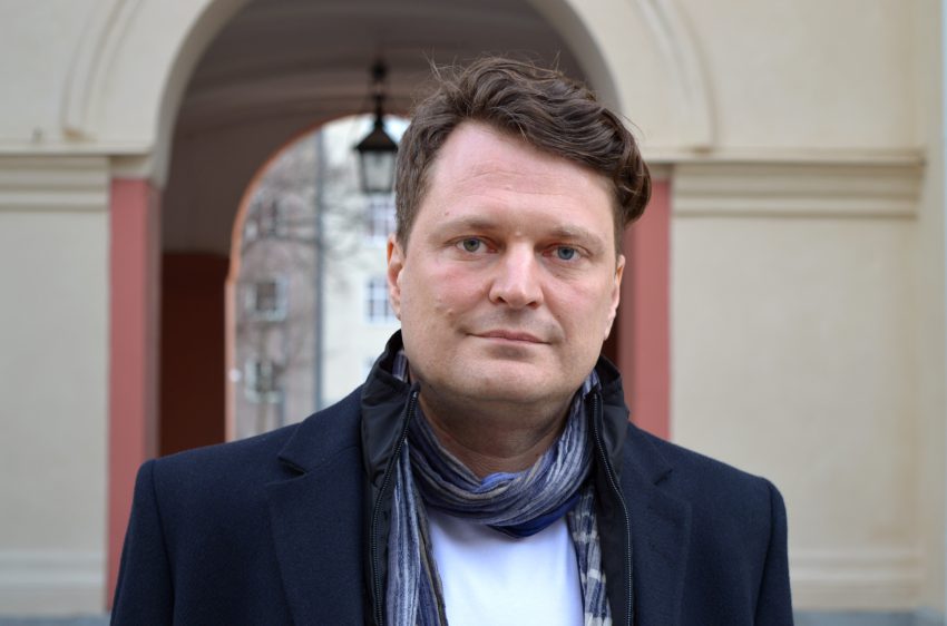 Daniel Velasco tittar in i kameran, utomhus klädd i rock och halsduk.